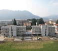 Newly constructed multi-story building, solar panels on the roof, swimming pool in front, surrounded by trees and grass