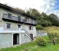2 story house, gray exterior, wooden balcony, stairs leading to the entrance, surrounded by grass and plants