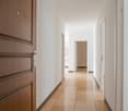 wide hallway, light brown tiles, white walls, wooden door, closed doors, corner room, open entrance