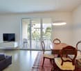 Spacious open-plan living and dining area with large windows providing a view of the outdoor area. The living room features a TV stand and a sofa, while the dining area has a wooden table with chairs. The room has a modern and minimalist design with a pen