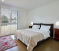 Spacious bedroom with large windows, hardwood floors, a king-size bed with a beige comforter, and a colorful patterned rug on the floor. The room has a modern and minimalist design with wooden nightstands on either side of the bed.