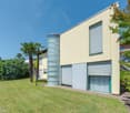 Modern 2 story house, yellow exterior, 2 garage doors, glass tower, greenery and palm trees in the surroundings