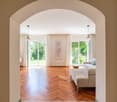 Spacious living room with large windows overlooking a garden, hardwood herringbone floor, white walls, arched entryway, pendant lighting, and a comfortable sofa