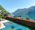Luxury villa with a swimming pool overlooking a scenic lake surrounded by mountains. The property has a well-manicured garden with a hedge lining the edge, providing privacy. The lake and mountains in the background create a picturesque and serene setting
