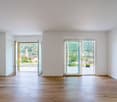 Spacious living room with large windows offering a scenic view of the surrounding landscape, including a church steeple in the distance. The room has hardwood floors and white walls, creating a bright and airy atmosphere.