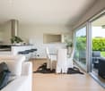 Open-concept living room and kitchen with large windows overlooking a garden, glass dining table with white chairs, beige sofa, and black rattan furniture on the patio outside