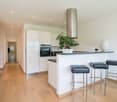 Spacious, modern kitchen with white cabinets, stainless steel appliances including a dishwasher, and a central island with bar stools. The kitchen opens up to a hallway and has hardwood floors throughout.