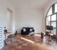 Spacious living room with large arched window, hardwood tile flooring, black sofa, desk with laptop, and music equipment