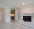 Spacious open-plan living area with white walls, marble-style tile flooring, recessed lighting, and a built-in TV cabinet. The kitchen area is visible through an archway, featuring white cabinetry and a built-in oven or dishwasher.