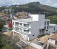 Modern white building, several floors, glass balconies, construction site in front, surrounded by houses and a mountain