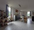 Dining area with wooden table, white chairs, white tiled floor, large window with curtain, white walls, framed picture, cat tower, potted plant, wooden shelves with books and picture frames, TV, white radiator, white desk with bowl, side table with plant,