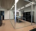 modern office room with glass walls and partitions, beige floor, table and chairs, black storage units, 3 paintings on the wall