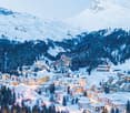 Mountain village covered in snow, alpine architecture, ski slopes, pine trees, large mountain range in the background.