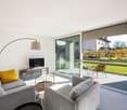modern living room with sliding glass doors, large windows, gray couches, glass table, yellow pillows, TV, floor lamp, balcony with chairs and table