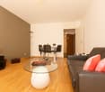 Spacious living room with hardwood floors, brown accent wall, glass coffee table, leather sofa, dining table with chairs, and a potted plant decoration