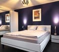 Spacious bedroom with a modern, minimalist design. The room features a large bed with white bedding, a built-in wardrobe, and a decorative lighting fixture. The walls are painted in a deep purple color, creating a cozy and sophisticated atmosphere. A fram