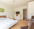 Spacious bedroom with hardwood flooring, white walls, a large bed with white bedding, a wooden headboard, a TV stand with a flat-screen TV, a comfortable armchair, and a small table with some decor items.