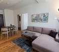 Spacious living room with hardwood floors, gray sofa, coffee table, and dining area with a round table and chairs. The room is decorated with tropical-themed artwork on the walls and has a floor lamp and air conditioning unit. There appears to be a balcon