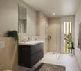 The bathroom has a white ceramic sink, a large mirror, and a floor-mounted shower with glass enclosure. A black cabinet with two drawers is next to the sink. The floor is tiled with white tiles, and there is a grey rug on the floor. The walls are tiled wi