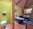 Bathtub, wooden beams, yellow wall, brick wall, mirror, shelves, toilet, counter