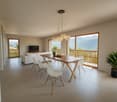 Spacious open-plan living and dining area with large windows offering a scenic mountain view. The room features a wooden dining table with white chairs, a white sofa, and a modern chandelier lighting fixture. The space has a clean, minimalist design with