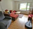 Spacious living room with large windows, hardwood floors, gray recliner sofa, red and yellow accent pillows, wooden coffee table, and shelving unit with decor items
