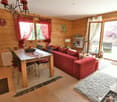 Cozy living room with wooden walls, red curtains, a large red sofa, a wooden dining table, and a view of the outdoors through the sliding glass door.