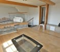 open plan living/sleeping area, hardwood floors, living room sofa with cushions, bed with bedding, white walls, kitchen with counter, window, balcony with railing, glass door to stairs, stool, wooden cabinet with drawers, and a black vase