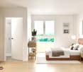 Bright and modern room with a bed, white bedding, pillows, and a bedspread. A side table holds books and a plant. A wardrobe with clothes, shoes, and bags. White flooring and walls, with a large window providing a scenic view.