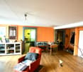 Living room with wooden floor, orange walls, white ceiling, wooden table, orange chair, white cabinet, picture frames, and various items on the table and cabinet.