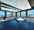 Modern office space with large windows offering a scenic view, a round meeting table with blue chairs, and a desk with a laptop and other office equipment.