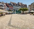 Cobblestone square with trees, multiple dining areas under white umbrellas, streetlight, groups of people sitting around tables, traditional European architecture, greenery