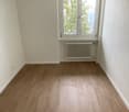 Empty bedroom with white walls, hardwood flooring, and a window overlooking trees outside.