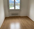 Empty room with a large window, wooden floor, radiator, and white walls