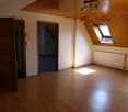 Spacious attic room with wooden paneled ceiling, hardwood flooring, and a large window providing natural light. The room has two doorways leading to other areas.