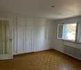 Spacious bedroom with white walls, hardwood parquet flooring, and built-in closets. The room has a large window providing natural light.