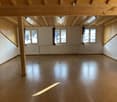 Large open room with wooden beamed ceiling, white walls, and hardwood flooring. The room has multiple windows providing natural light. The space appears to be empty and suitable for various uses.