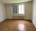 Spacious bedroom with hardwood parquet flooring, white walls, and a large window with shutters. The room appears to be empty, ready for furnishing.