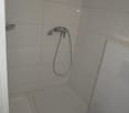 Small shower area, white tiles, toilet on the right, silver shower head, flush button on the wall