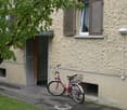 Beige exterior wall, red bicycle parked in front, small grassy area, sidewalk, tree