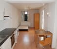 Open-plan studio apartment with kitchen, dining area, and living space. Hardwood floors, white walls, kitchen appliances including a dishwasher, and a wooden cabinet.