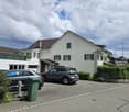 2-story white house with brown roof, windows with green shutters, 2 cars parked in front
