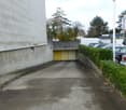 An empty parking lot leads to a yellow garage entrance. There are several cars parked in front of a building. The parking lot is concrete with some dried leaves and has bushes on the side. There is a pole with the word Atlas on it.