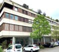 multistory building, glass windows, brown facade, several cars parked in front, shrubs and trees