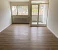 Spacious living room with hardwood flooring, large windows providing natural light, and a sliding glass door leading to a balcony or terrace with a view of the surrounding neighborhood.