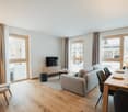 Spacious open-plan living room and dining area with large windows, hardwood floors, gray sofa, wooden TV stand, and dining table with chairs. The room has a cozy and modern feel.