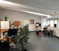 The image shows an open office space with a tiled floor, white walls, and exposed ceiling beams. The room is furnished with various office equipment and furniture, including desks, chairs, shelves, and a large potted plant. There is also a dishwasher visi