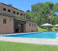 Building exterior with swimming pool, brick walls, tiled roof, outdoor seating, umbrellas, potted plants, green lawn, trees.