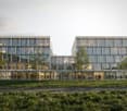 Modern office building with glass facade, multiple floors, entrance with glass doors, people walking around, trees, and grass in front.