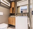 Modern bathroom with wooden vanity cabinet, white sink, mirror, and toilet. Shelving unit and towel rack visible.